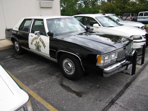 1991 Ford Crown Victoria - A police car with a push bar is parked in a lot next to other vehicles.
