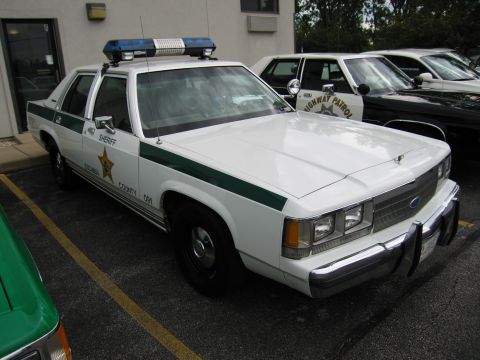 1991 Ford Crown Victoria - A white 1991 Ford Crown Victoria police car is parked next to other vehicles, including one marked "HIGHWAY PATROL".