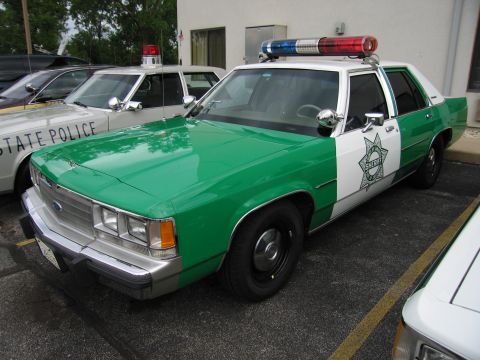 1991 Ford Crown Victoria - A 1991 Ford Crown Victoria police car, painted green and white with a sheriff's star on the door, is parked next to other vehicles.