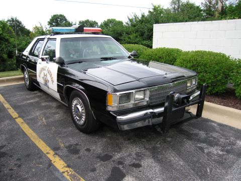 1991 Ford Crown Victoria - A black and white police car is parked in a lot, a push bumper and emergency lights.