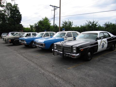 1991 Ford Crown Victoria - A line of vintage police cars, some blue and white, and a black and white patrol car, are parked in a lot.