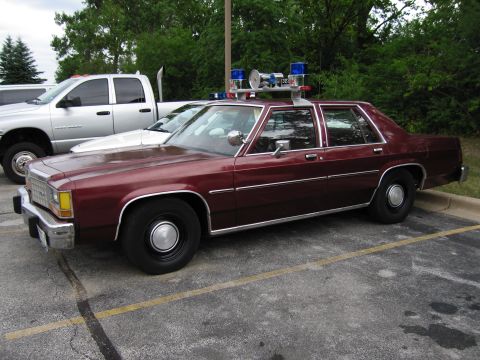 1991 Ford Crown Victoria - Side view of a maroon police car equipped with a light bar and siren, parked in a lot.