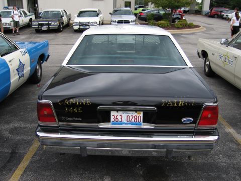 1991 Ford Crown Victoria - A 1991 Ford Crown Victoria police car is parked next to other vehicles, showing its rear end with police markings.