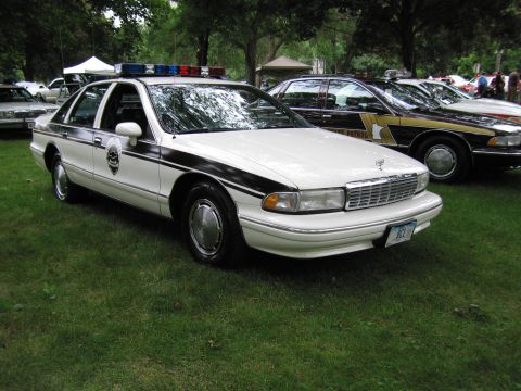 1991 Ford Crown Victoria - This white police car, possibly a 1991 Chevrolet Caprice, features a distinctive black stripe and a light bar on the roof.