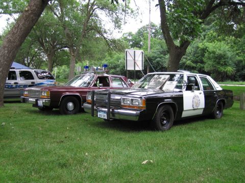 1991 Ford Crown Victoria - Two older model police sedans are parked on a grassy area with trees in the background.