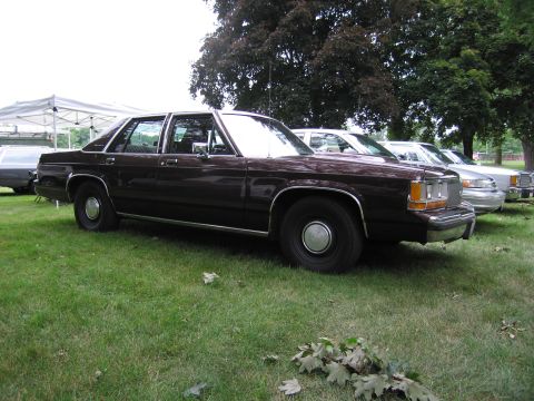 1991 Ford Crown Victoria - A dark brown sedan is parked on a grassy lawn next to other vehicles under trees.