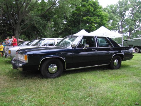 1991 Ford Crown Victoria - A dark blue sedan is parked on a grassy area with trees and tents in the background.