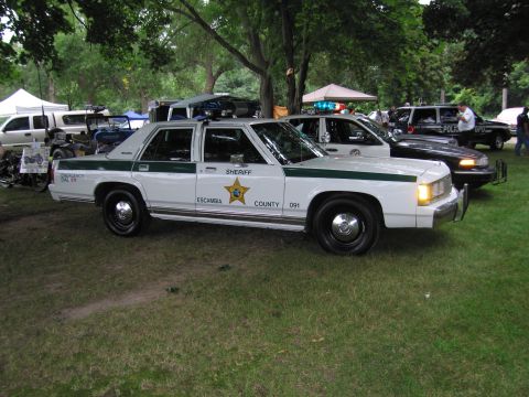 1991 Ford Crown Victoria - A white 1991 Ford Crown Victoria police car is parked on grass with other vehicles and tents in the background.
