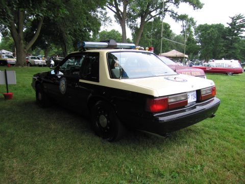 1991 Ford Crown Victoria - A black and tan police car is parked on the grass with other vehicles in the background.