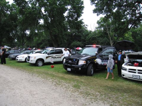 1991 Ford Crown Victoria - A lineup of police vehicles, including SUVs and cars, are parked on a grassy area under a cloudy sky.