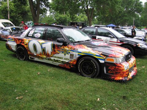 1991 Ford Crown Victoria - A heavily customized police car with flame graphics and "01" livery is parked on grass next to other vehicles.