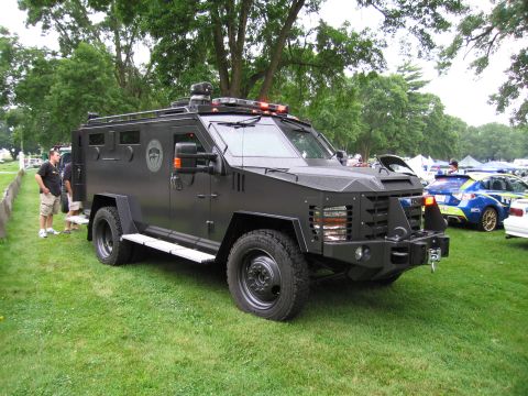1991 Ford Crown Victoria - An imposing armored vehicle sits on display on a grassy field, with other vehicles visible in the background.