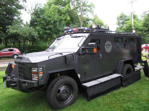 1991 Ford Crown Victoria - A black armored truck with emergency lights is parked on a grassy area next to a road.