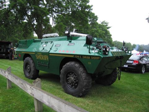 1991 Ford Crown Victoria - A green armored vehicle, marked as 'SHERIFF', is parked on a grassy field next to a police car.