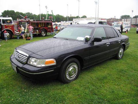 1991 Ford Crown Victoria - A dark sedan sits on a grassy field with emergency vehicles and tents in the background.