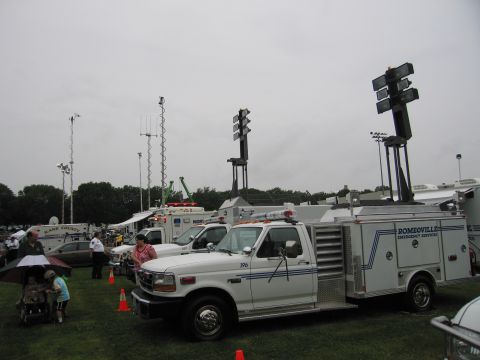 1991 Ford Crown Victoria - A white emergency service truck with a light tower is parked on grass, with other emergency vehicles and people in the background.