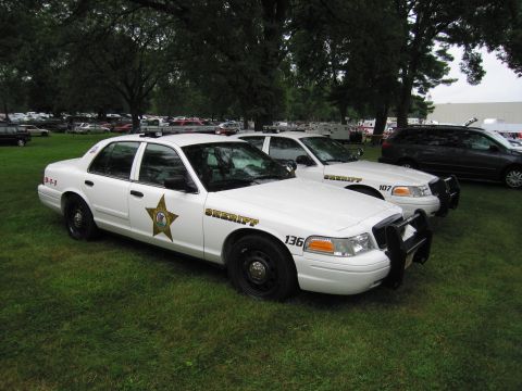 1991 Ford Crown Victoria - Two white sheriff's cars are parked on a grassy field, with a dark SUV in the background.