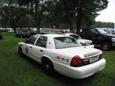 1991 Ford Crown Victoria - A white sheriff car is parked on a grassy field, with other vehicles and trees in the background.