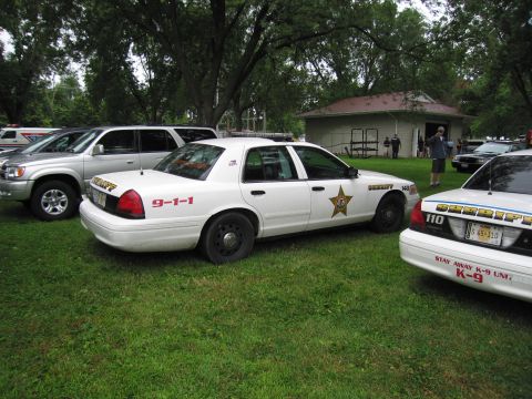 1991 Ford Crown Victoria - A white police car with a sheriff star and 9-1-1 marking is parked on the grass next to other vehicles.