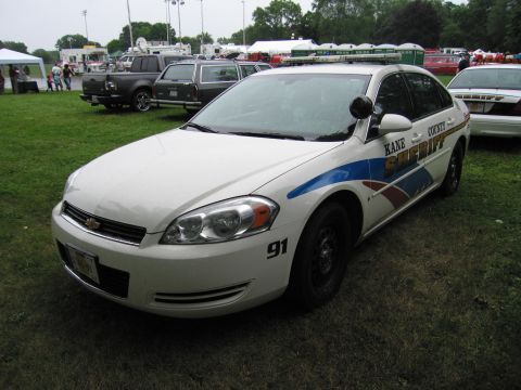 1991 Ford Crown Victoria - A white police car with "KANE COUNTY SHERIFF" markings is parked on grass, with other vehicles and tents in the background.