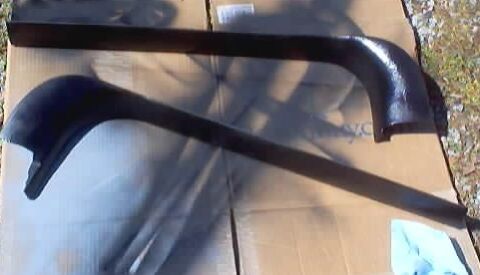 1999 Ford Taurus - A pair of long curved black interior trim moldings, likely door sill or rocker/kick panel pieces, laid out on flattened cardboard outdoors…