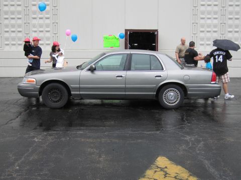 1991 Ford Crown Victoria - A silver sedan is parked on a wet asphalt surface with people and balloons around it.