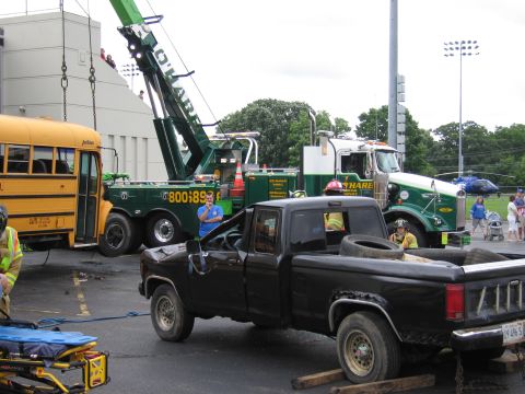 1991 Ford Crown Victoria - A tow truck is lifting a school bus, while a damaged black pickup truck sits in the foreground, with a helicopter in the background.