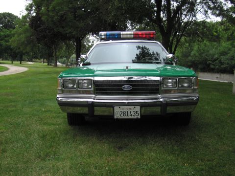 1991 Ford Crown Victoria - A green 1991 Ford Crown Victoria police car is parked on a grassy area with trees in the background.
