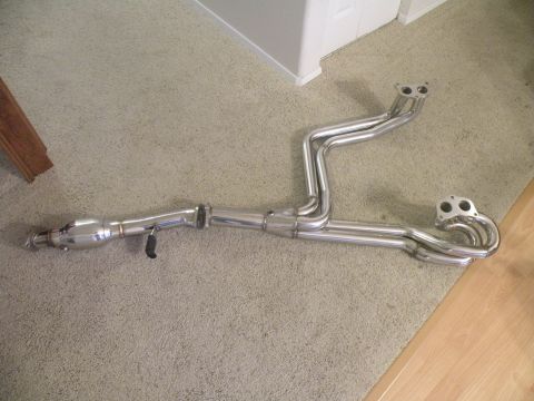 1998 Subaru Legacy - A shiny metal exhaust manifold with a catalytic converter and dual exhaust pipes is displayed on a carpeted floor.