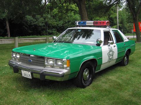 1991 Ford Crown Victoria - A police car with a sheriff's star emblem on the door sits on a grassy lawn.
