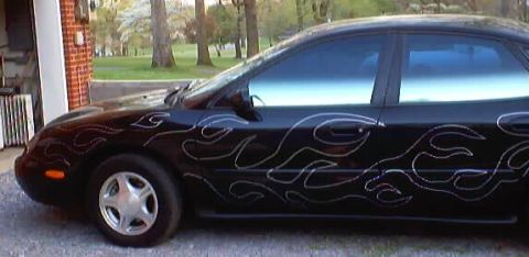 1999 Ford Taurus - A black 1999 Ford Taurus sedan is visible from the side, adorned with silver flame decals.
