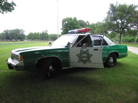 1991 Ford Crown Victoria - A vintage police car, possibly from the 1980s, is parked on a grassy area with trees in the background.