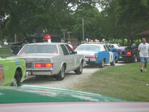 1991 Ford Crown Victoria - Several police cars are lined up on a gravel road, with trees and a building in the background.