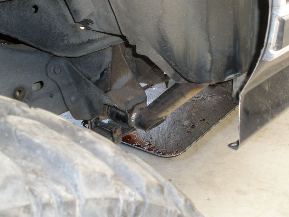 Undercarriage view showing suspension components, exhaust pipe, and a large tire. Some fluid is visible on the ground
