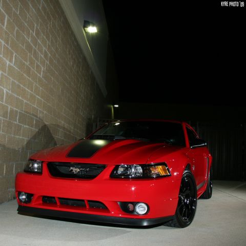 2004 Ford Mustang - A red 2004 Ford Mustang is parked in front of a brick wall at night, its distinctive front grille and black hood stripe.