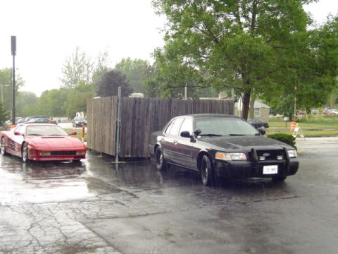 2000 Ford Crown Victoria - Two cars, a police interceptor and a red sports car, are parked outside on a rainy day.