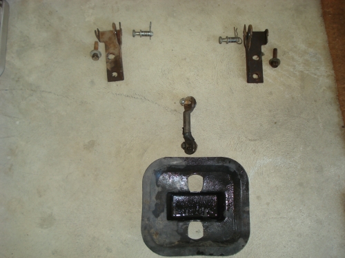 Several metal vehicle parts, including brackets, bolts, and a rubber-coated plate, laid out on a concrete surface