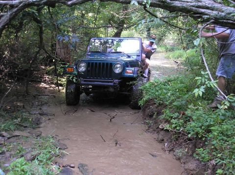 1987 Suzuki Samurai - Head-on view of a dark green Jeep Wrangler TJ wading through a muddy creek crossing in dense woods, running round headlights, a tube front…