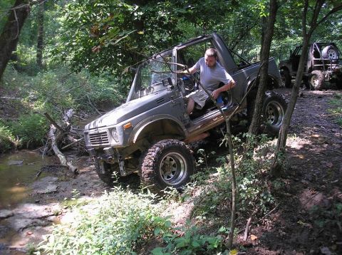 1987 Suzuki Samurai - A lifted olive-drab doorless mini 4x4 with a bare roll cage and no top crawls down a muddy creek bank in the woods, front wheels dropping t…