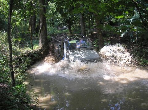 1987 Suzuki Samurai - A tan topless Suzuki Samurai 4x4 with a roll bar and folded-down windshield fords a shallow wooded creek, throwing up large splashes of wat…