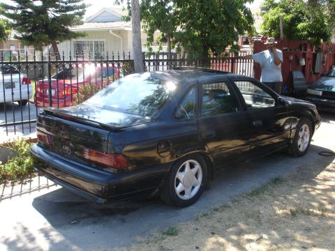 1991 Ford Taurus - Rear view of a black 1991 Ford Taurus sedan parked on a driveway with a house and fence in the background.