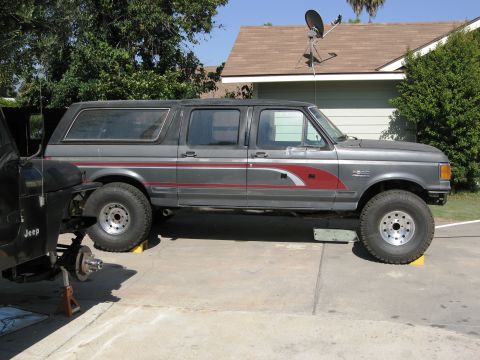 1986 Ford Bronco - Gray Ford Bronco SUV with red stripe, lifted on yellow blocks, side view, with a Jeep in the foreground.