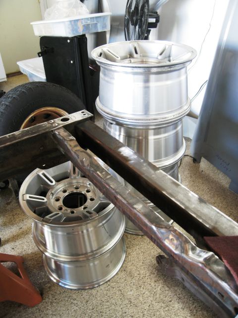1986 Ford Bronco - Several aluminum wheels are stacked next to a tire and some metal framing, suggesting parts for a vehicle restoration.