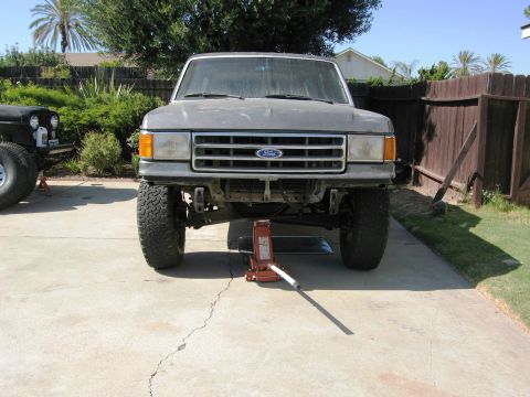 1986 Ford Bronco - A gray Ford Bronco is jacked up in a driveway, with another vehicle to its left.