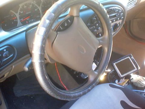 1999 Ford Taurus - Driver view of a tan Ford sedan interior with cloth-wrapped steering wheel, gauges, and a mounted screen.