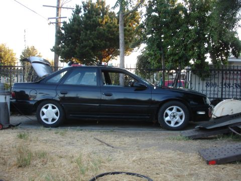 1991 Ford Taurus - Side profile of a black 1991 Ford Taurus with the trunk open, parked on dry grass.