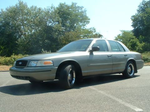 2000 Ford Crown Victoria - A tan 2000 Ford Crown Victoria is shown from the front three-quarter angle with trees and a clear sky behind it.