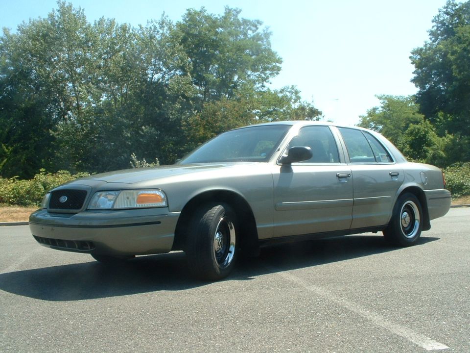 Tan 2000 Ford Crown Victoria sedan parked on asphalt, front three-quarter view, trees in background