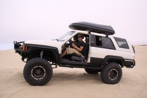 1985 Toyota 4Runner - A modified SUV with oversized tires and a cargo box on the roof sits on a sandy terrain.