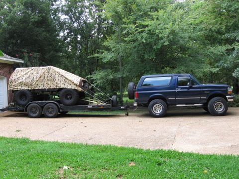 1967 Ford Mustang - A blue Ford Bronco is hitched to a trailer carrying a large, covered object with multiple wheels.